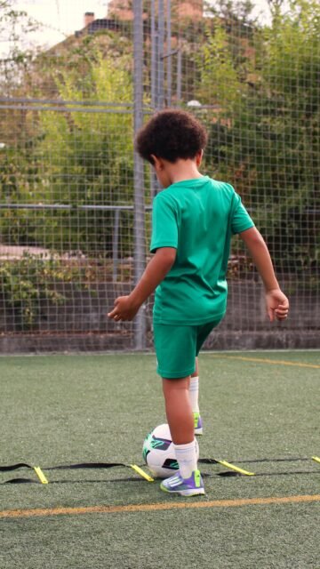 Niño pequeño conduciendo el balón junto a una escalera de agilidad en el campo de fútbol de San Blas