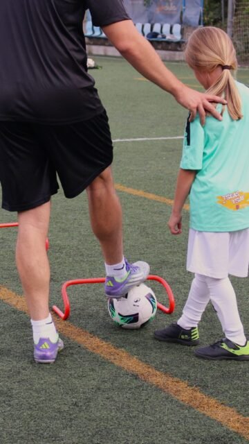 Entrenador y niña practicando el control del balón junto a una valla de agilidad en la escuela High Tech Fútbol